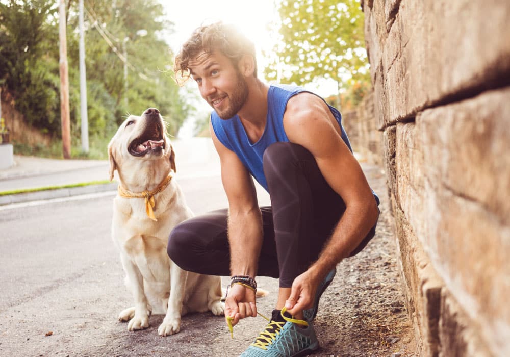 A man tying his shoes, he has a dog sitting next to him. The man is also wearing running clothes.