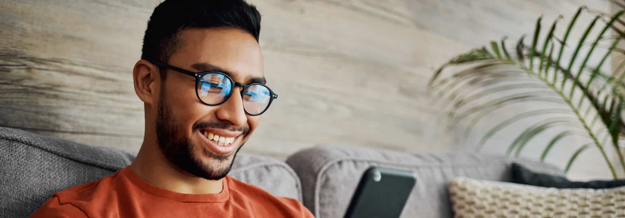 A man looking at his cell phone while sitting on the couch. He is smiling and wearing glasses.