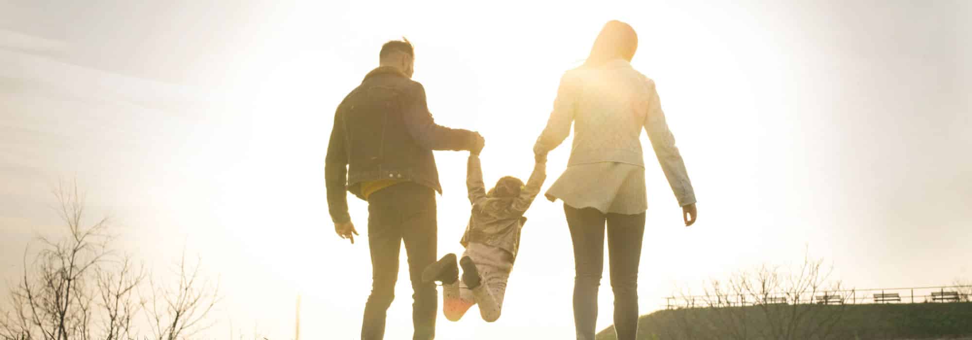 A family appearing to be on a walk. The couple has a child between them, they are each holding the child's hand and swinging them back and forth.