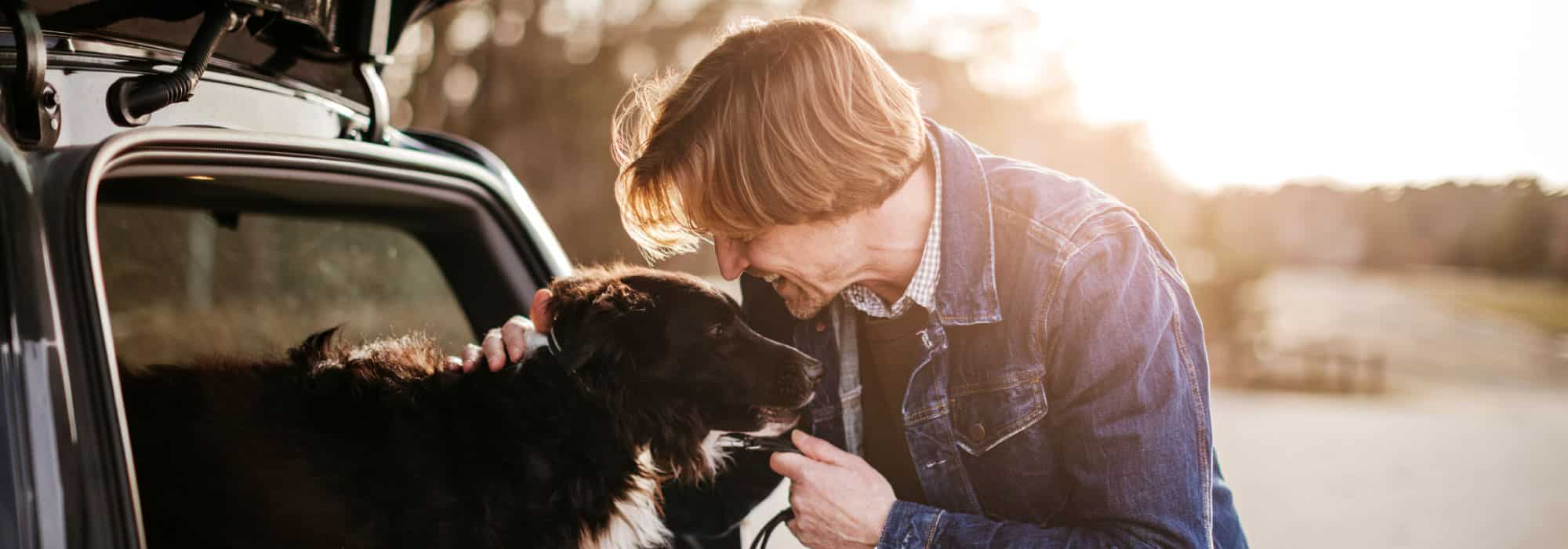 A man petting a dog, the dog has its' head outside of the window of a car. The man is smiling.
