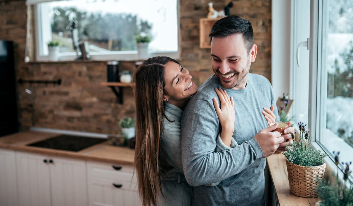 Woman hugs man from behind while they're laughing and standing in the kitchen by a window