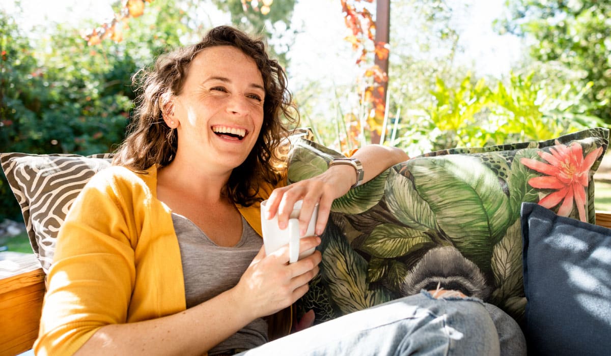 Woman laughs while holding a coffee mug and sits on a couch outside.