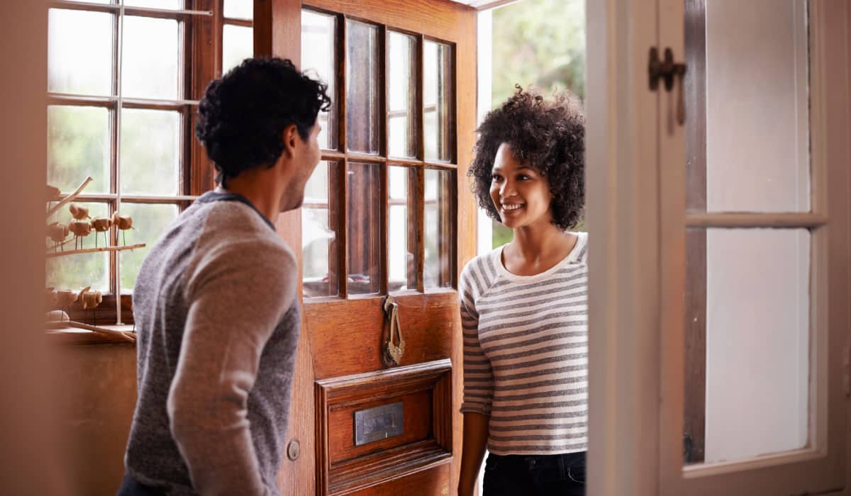 A man opening a front door to a house and speaking to a woman who is coming inside.