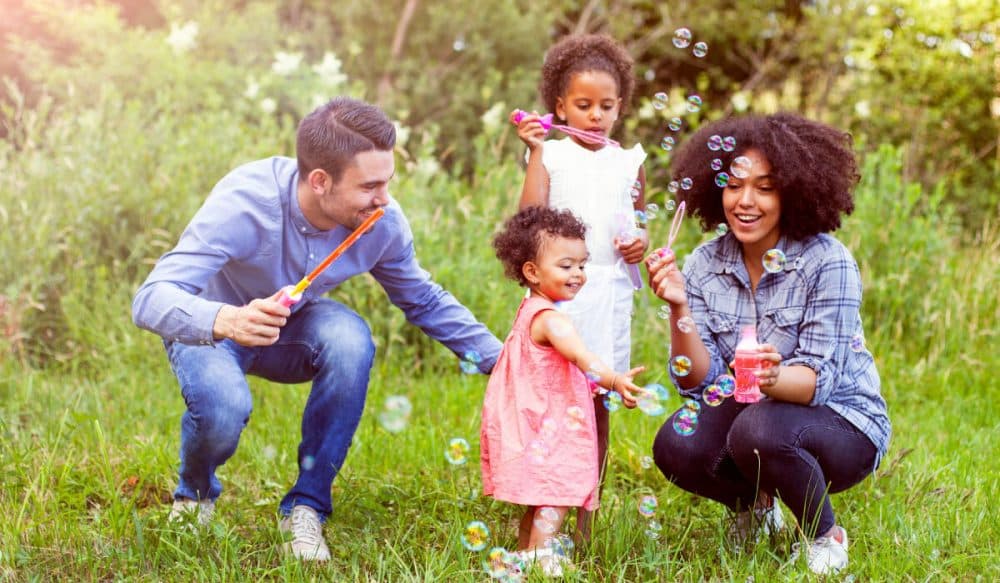A family blowing bubbles. They are laughing and appear to be at a park. There is a man, a woman and two little girls.