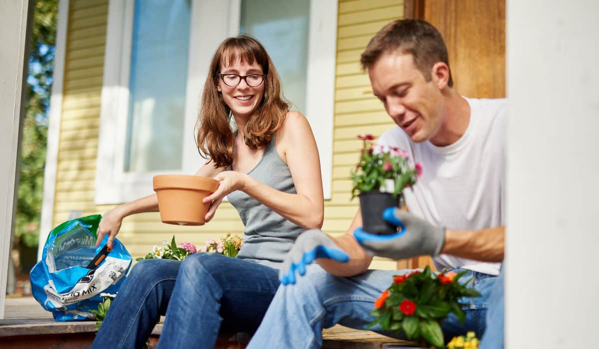 A couple sitting on the front porch planting flowers in pots. He is wearing garden gloves and she is holding a pot.