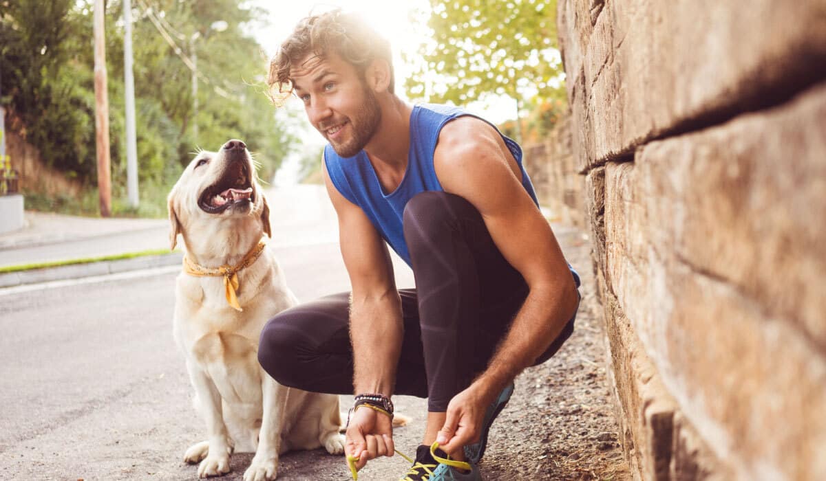 A man tying his shoes, he has a dog sitting next to him. The man is also wearing running clothes.
