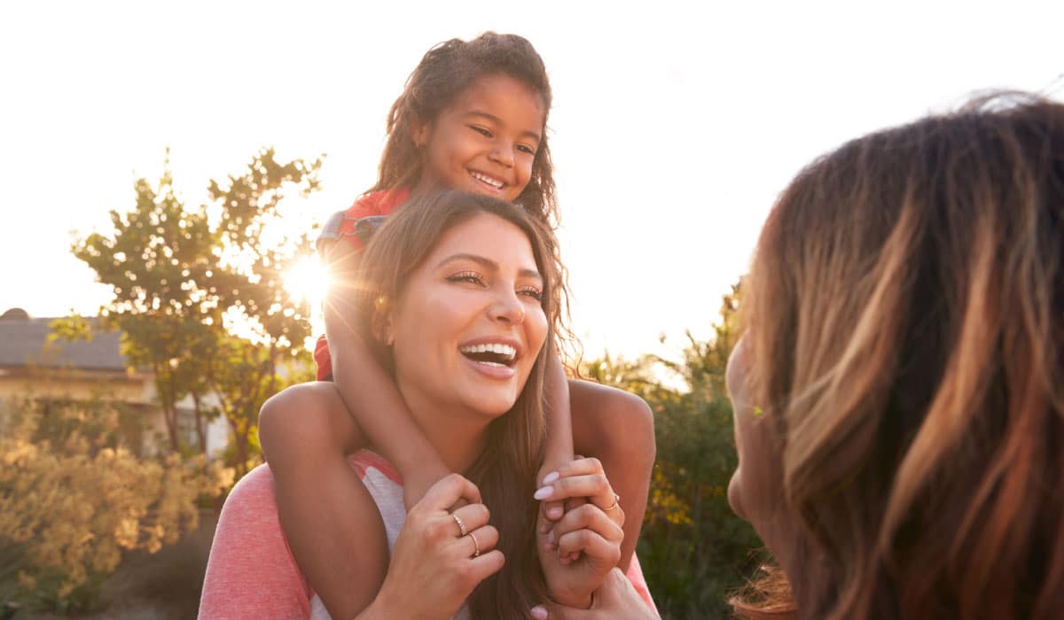 A woman carrying a child on her back and speaking to another woman. They are laughing.