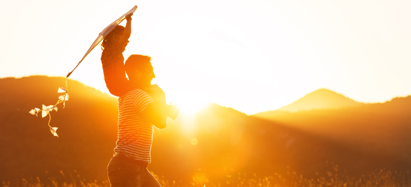 Child with a kite on a man's shoulders walking in front of a sunset between hills.