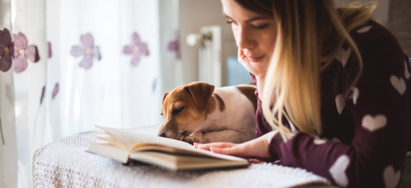 A woman laying on a bed reading a book. A dog is laying next to her.