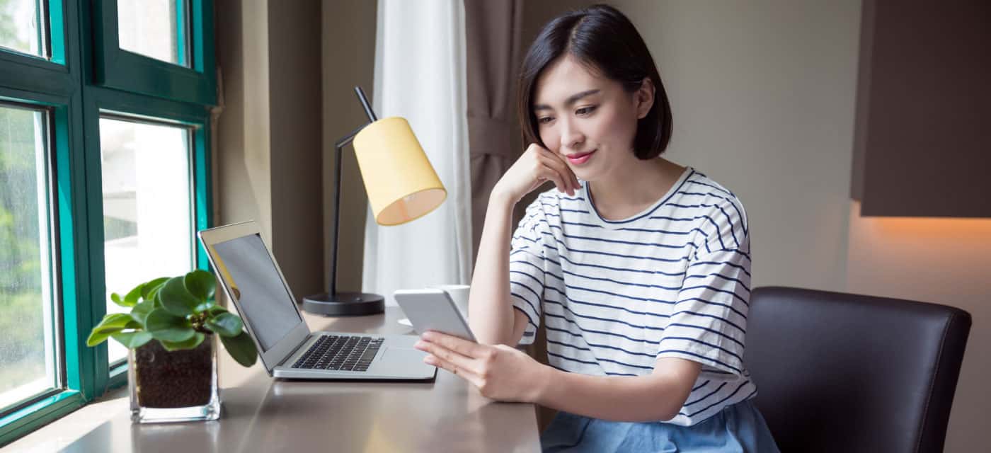 A woman sitting at a desk, there is an open laptop on the desk and she is using a cell phone.