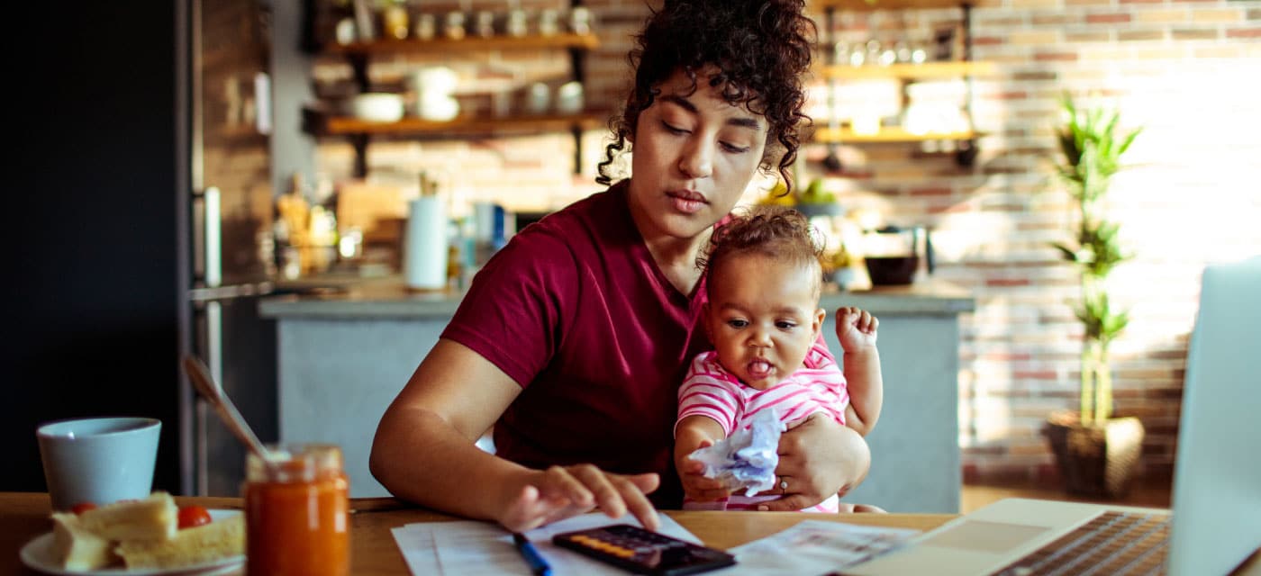 A woman sits at a table with a baby. She is using a calculator on her phone.