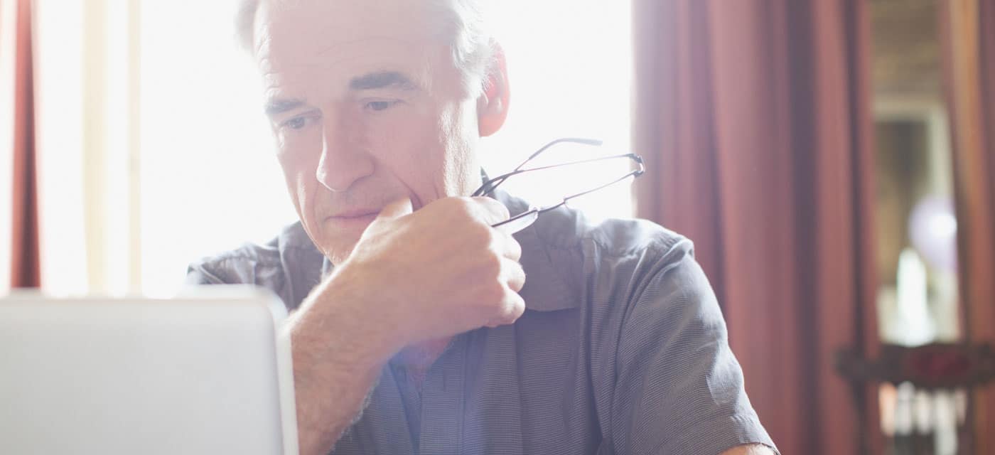 An older man looking at an open laptop, he is holding a pair of glasses in his hand.