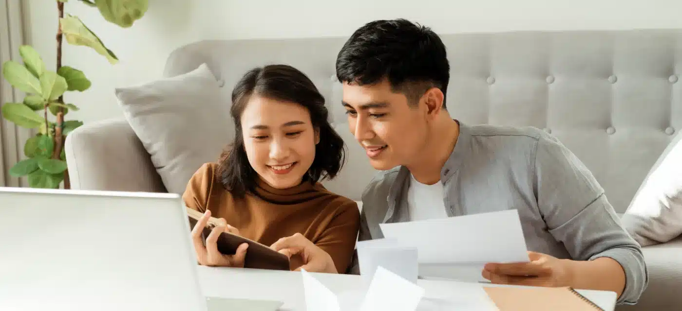 Couple sit on floor in front of couch looking at a notebook and paper with a laptop in front of them.