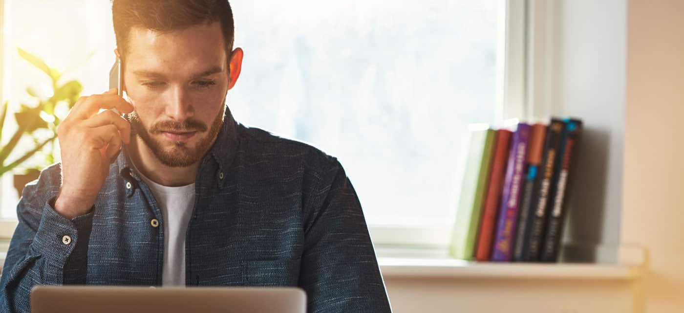 A man sitting at a table and using a laptop, he is on the phone.