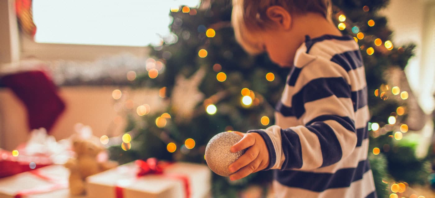 A child holding a Christmas ball and standing in front of a Christmas tree.