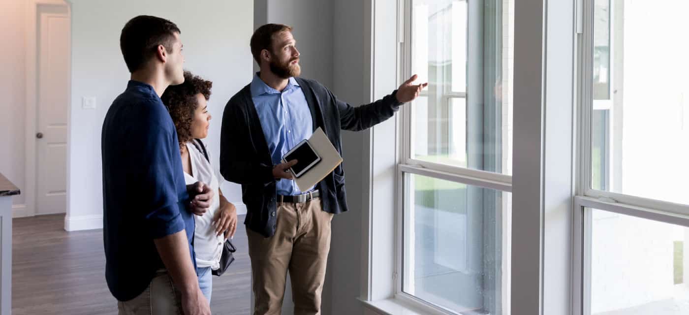 A couple walking through a house and standing in front of a window. They appear to be with a realtor.
