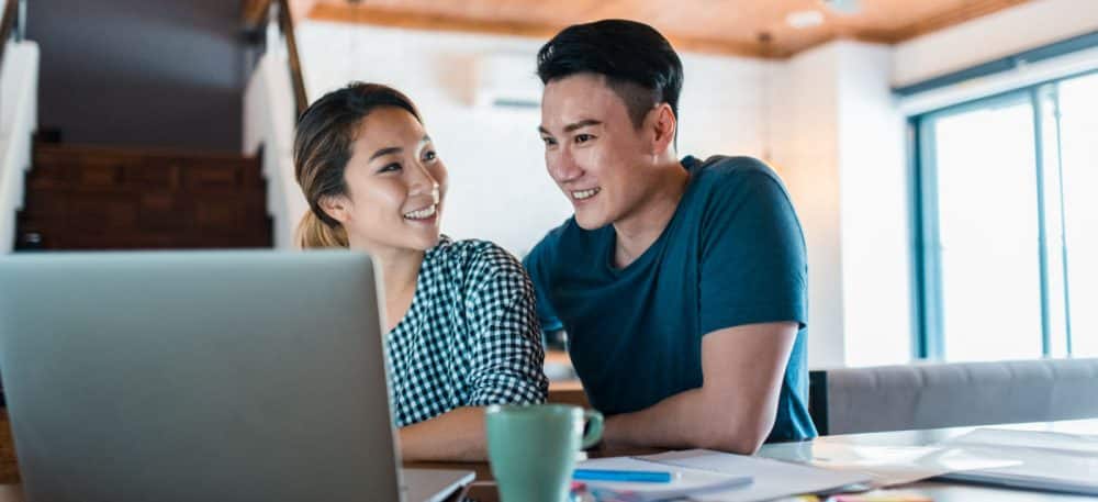 A couple sitting at a kitchen table with a laptop in front of it.