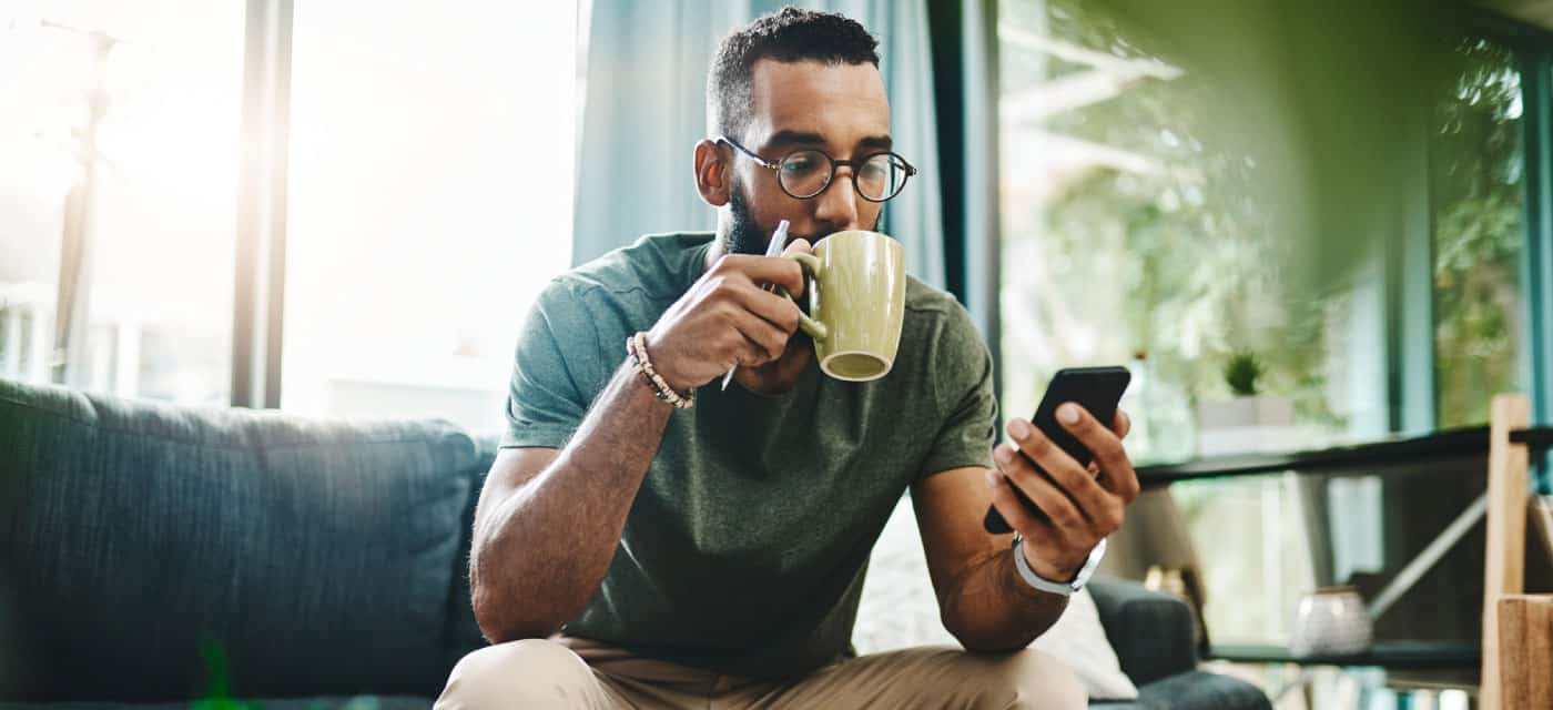 A man sitting on a couch, using his cell phone and drinking a cup of coffee