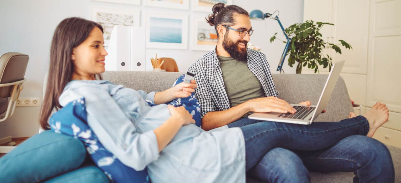 A couple sitting on a couch. The woman is holding a credit card and the man is holding an open laptop. They are smiling.