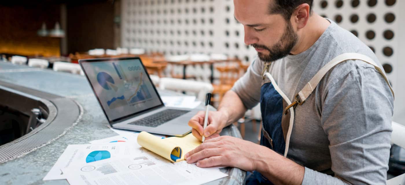 Man sitting at a table and writing on a notepad. He has a laptop in front of him with graphs on it.