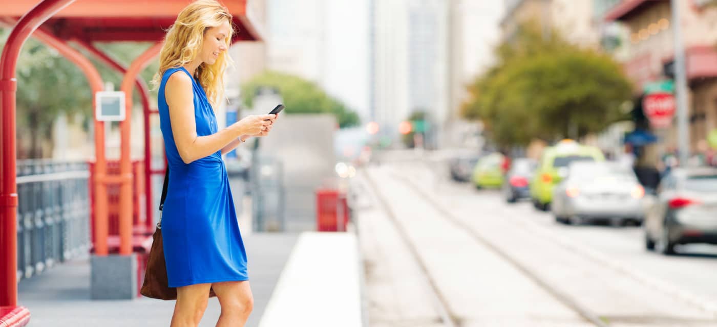 A woman standing outside by train tracks, she is using a phone and smiling.