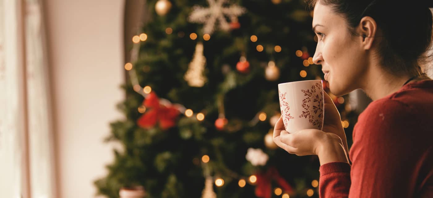 A woman sitting in front of a Christmas tree and holding a holiday mug.