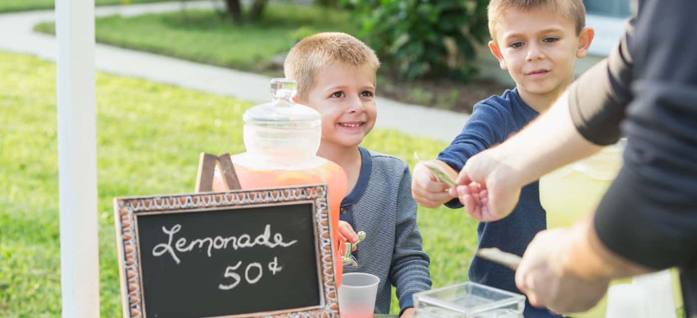 Two kids selling lemonade at a lemonade stand. An adult is giving them money.