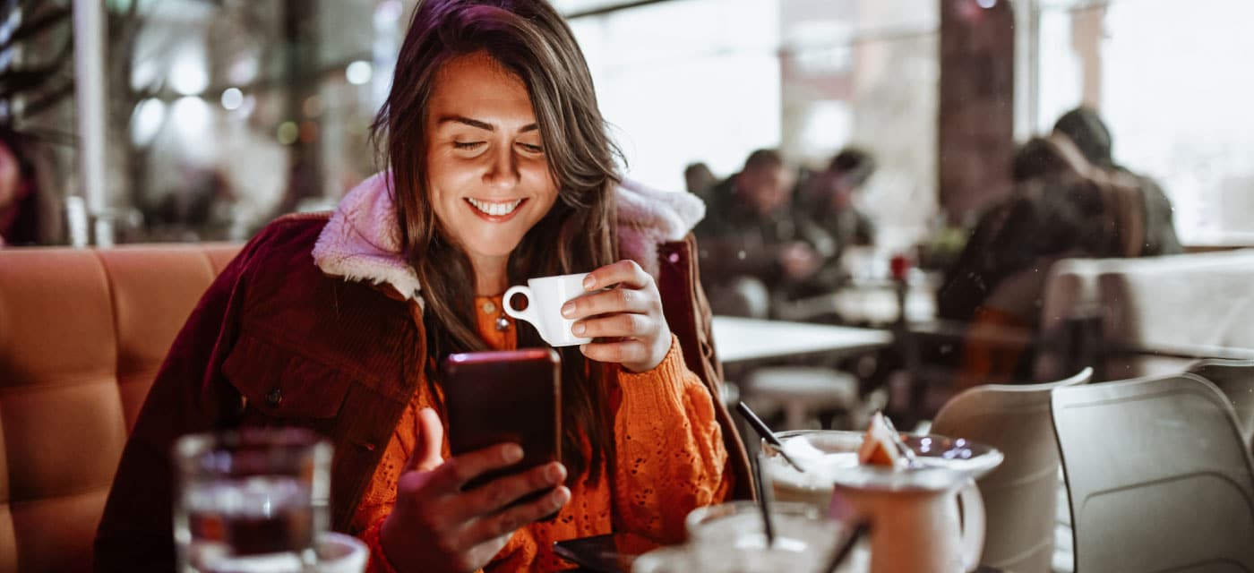 A woman sits at a table and sips a cup of coffee, she is using a cell phone and smiling.