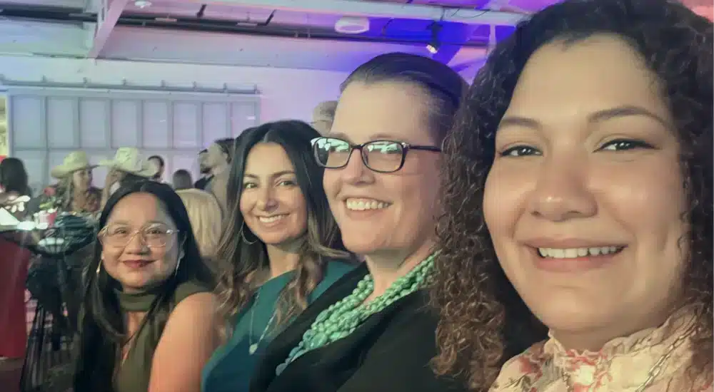 Group of smiling women posing for a selfie at a party with colorful lighting in the background.