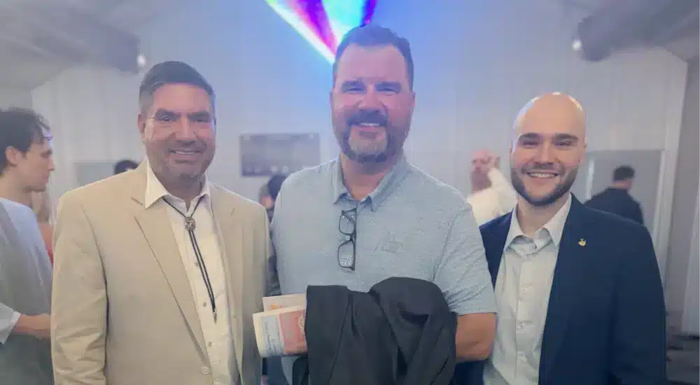 Three men in business attire pose and smile for a photo at an indoor event. The man in the middle holds a coat and some pamphlets.