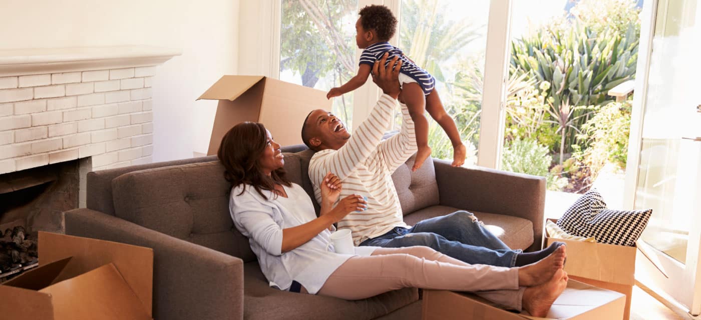 a family sitting on a couch with a baby, the man is tossing the baby in the air and smiling. There's moving boxes around them.