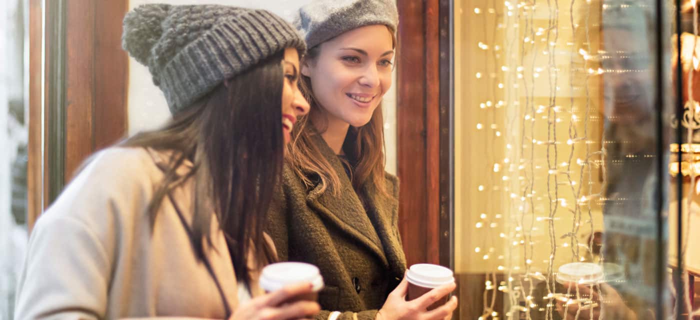 Two women looking in a store window, they are holding coffee and smiling.