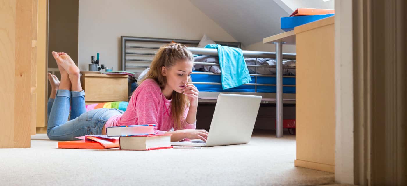 A woman laying on the floor of a dorm room, surrounded by books and using a laptop.