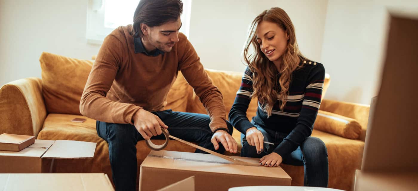 Couple sits on couch and tapes up moving box.