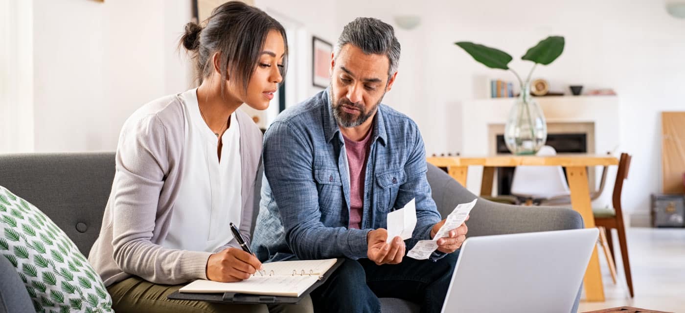 A couple sitting on a couch and looking at receipts, they are sitting in front of an open laptop.