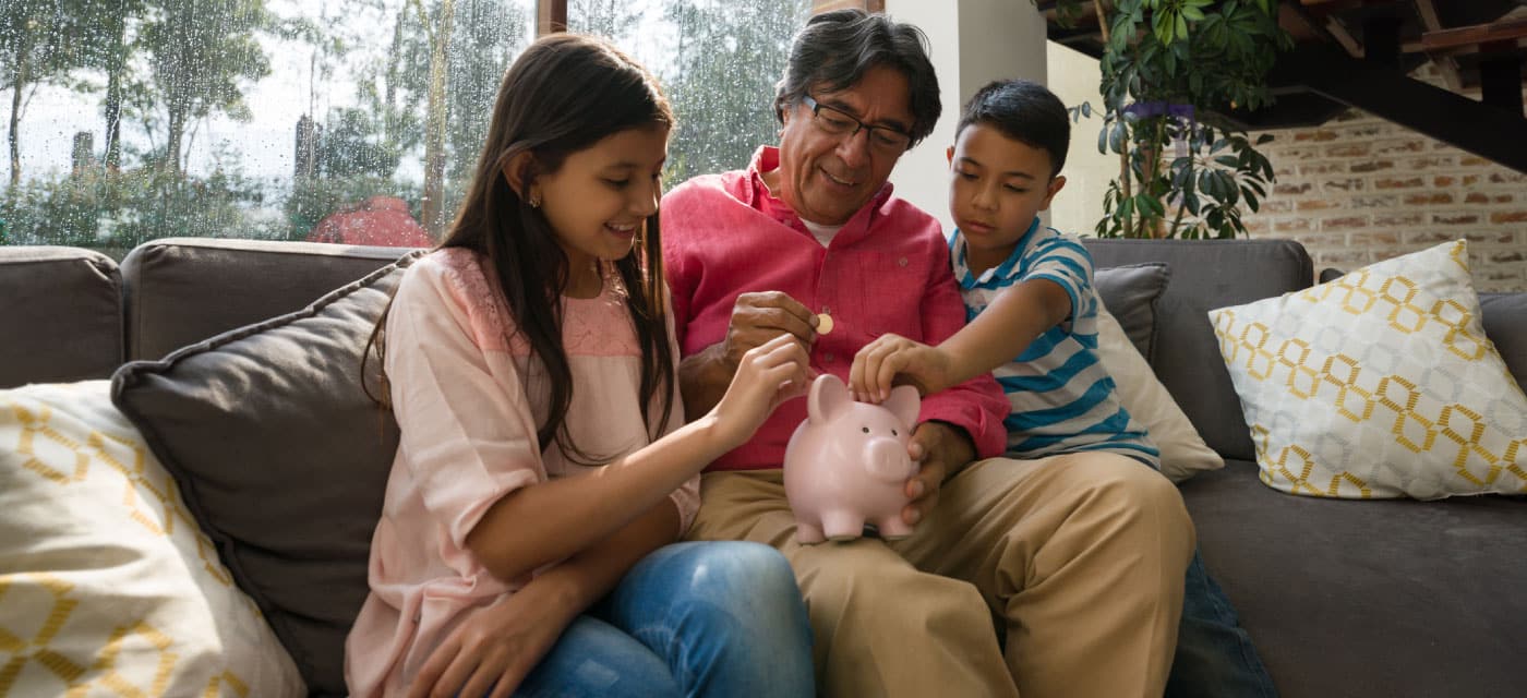 An older man and two young kids, they are holding a piggy bank between them and sitting on a couch.
