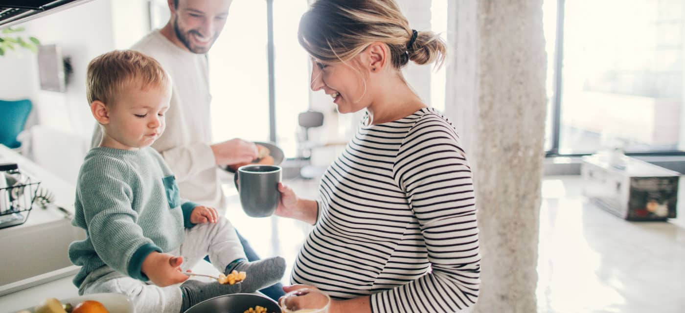 A couple feeding a baby. They are standing in the kitchen, the woman is holding a mug and is pregnant. The man is smiling.