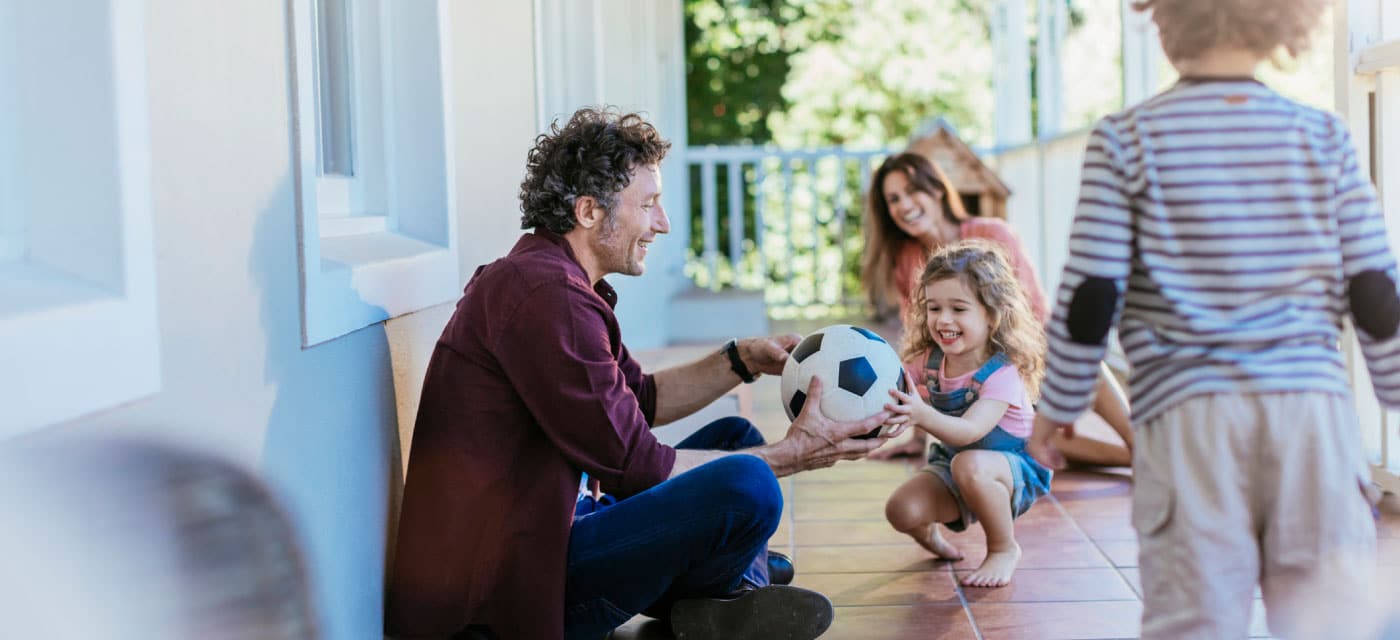 A family on a front porch, the man is holding a soccer ball and giving it to a little girl.