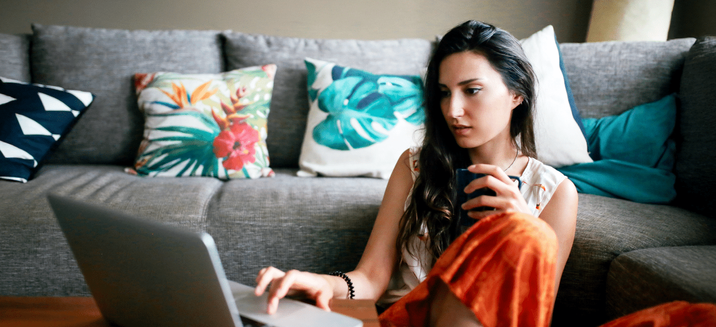 Woman sitting in front of her couch drinking coffee and studying her laptop.