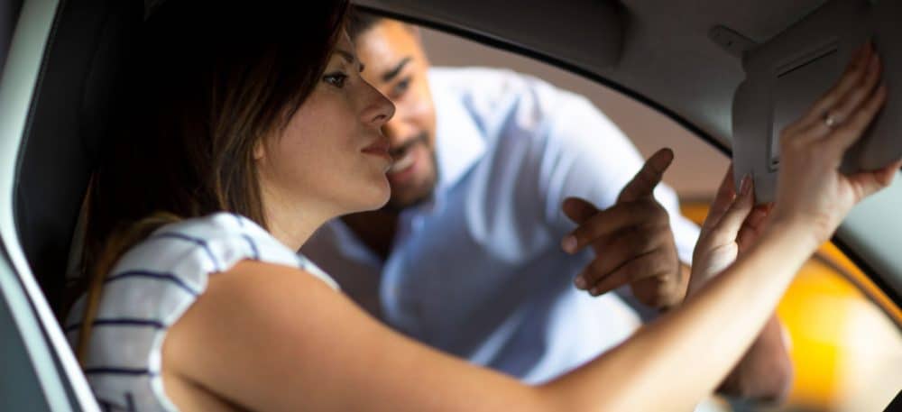 A woman sitting in a car and looking into the the mirrors, a man is standing outside the car and pointing at the mirror.