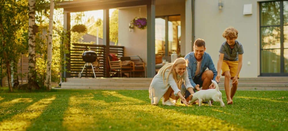 A family standing outside a house, in the yard, they are playing with a puppy and smiling.