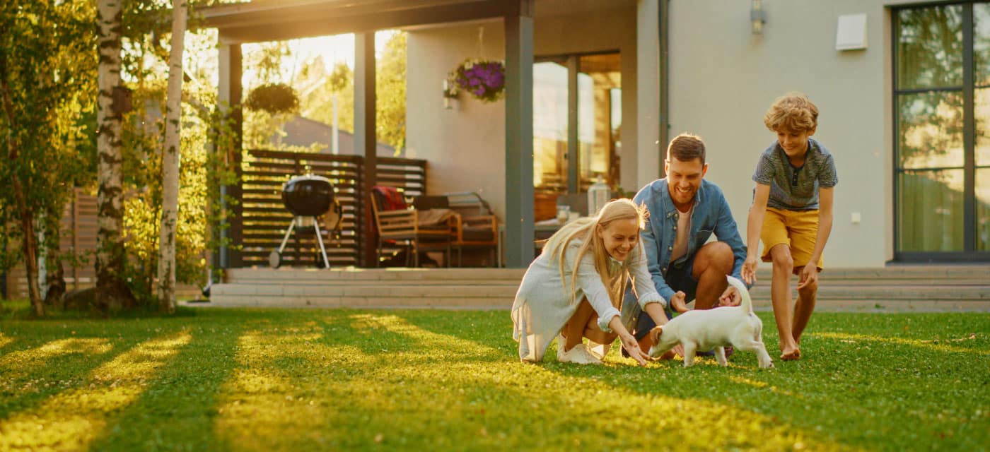 A family standing outside a house, in the yard, they are playing with a puppy and smiling.