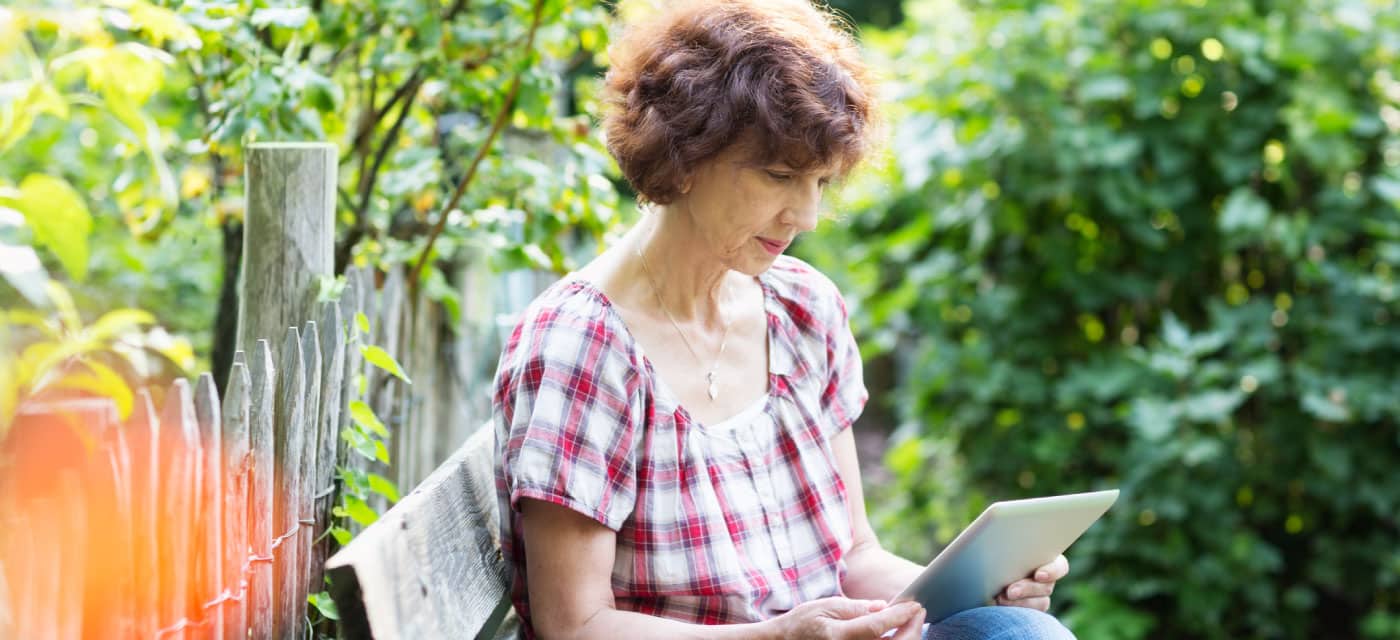 A woman reading on an IPad. She is sitting outside on a bench.