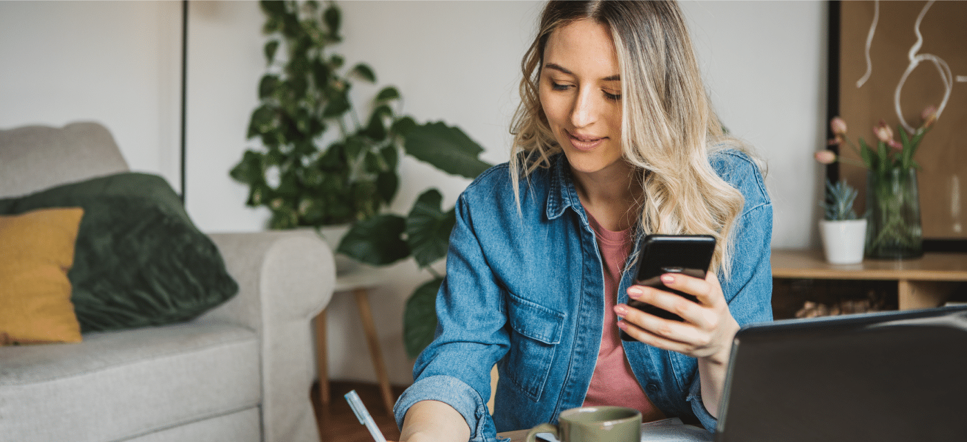 Woman in front of laptop holding smartphone writes something down.