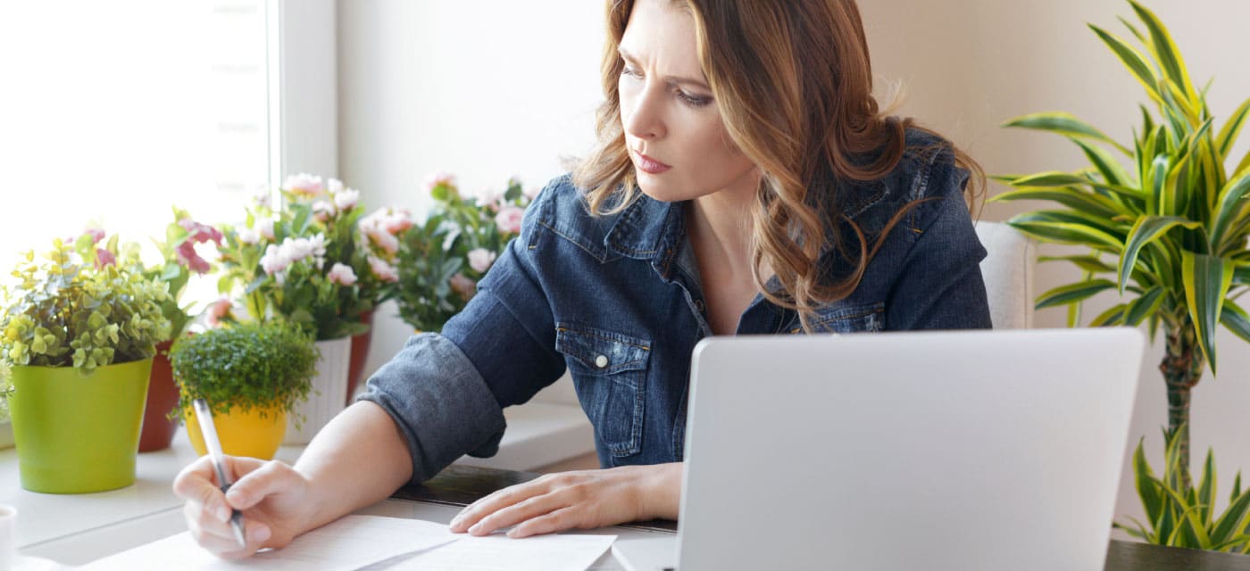 A woman sitting at a table with a laptop, she is writing on a piece of paper.