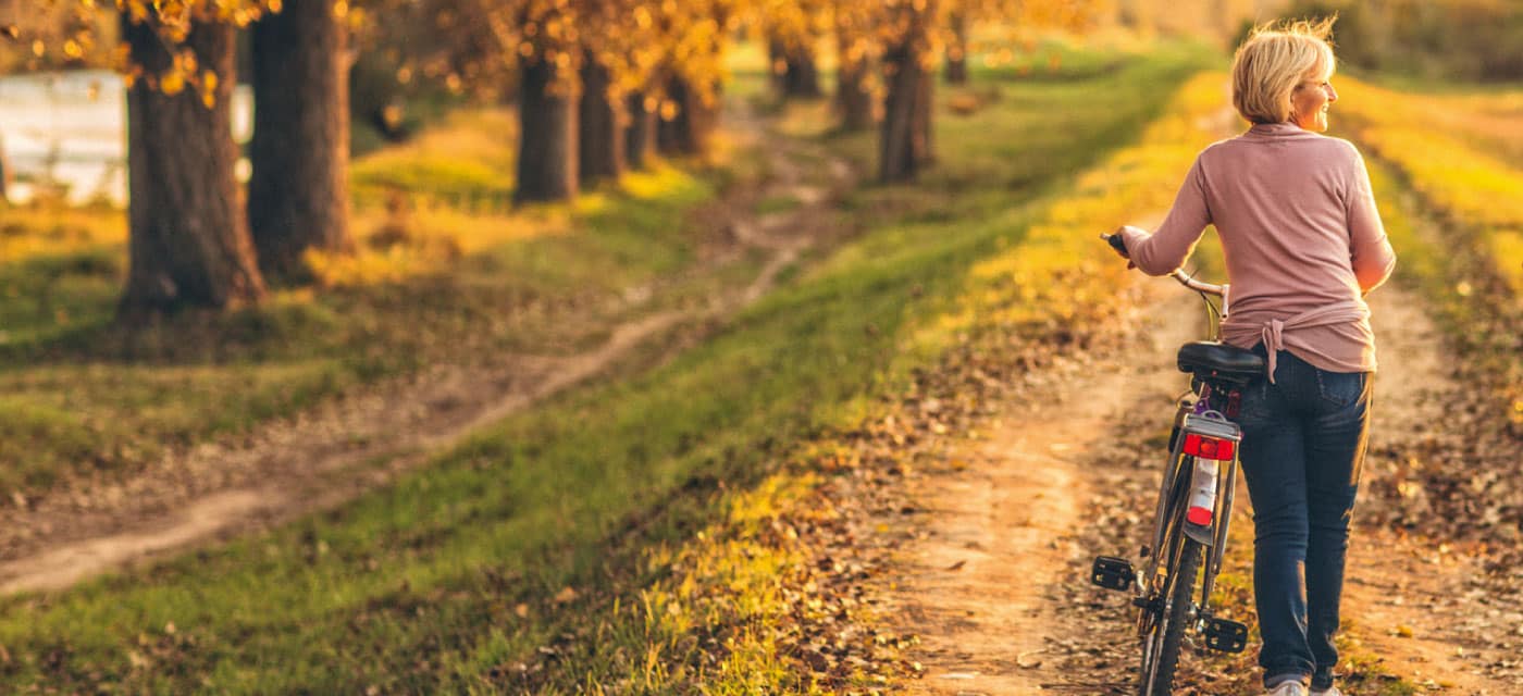 A woman is walking on a dirt path with a bike. It appears to be autumn.