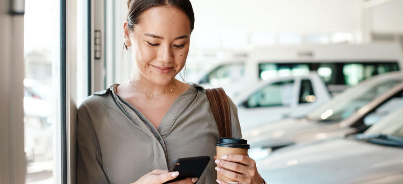 Woman stands against a wall in an auto dealership while holding a coffee and looking at her phone.