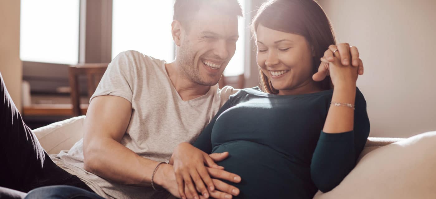 A couple sitting on a couch and smiling, the woman is pregnant and the man has his hand on her stomach.