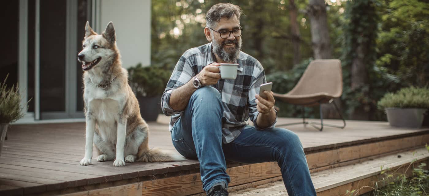 A man sitting on a porch with his dog. He is drinking out of a mug, smiling and looking at his phone.