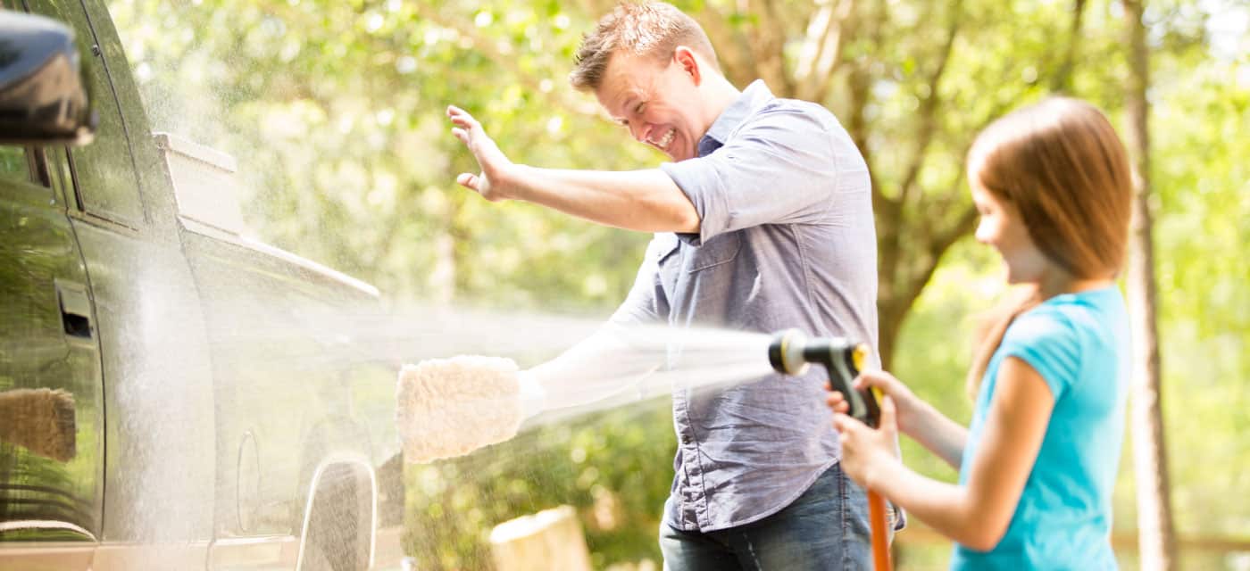 A man and a young girl washing a truck. The girl is using a strong hose and the man is laughing.